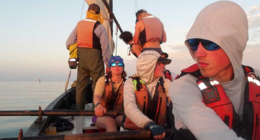 A group of students wearing lifejackets work on a sailboat. Some are holding oars while others work with the sail. 